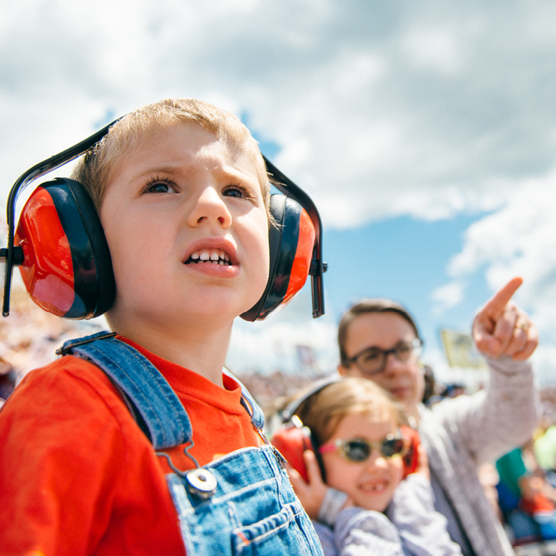 A young child wearing red ear protection headphones looks up with an excited expression while attending an outdoor racing event with family