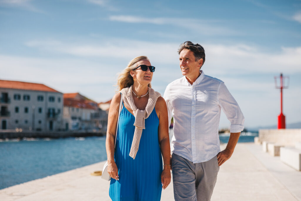 An older couple walks along a sunny coastal waterfront, smiling and enjoying the ocean breeze near a harbor