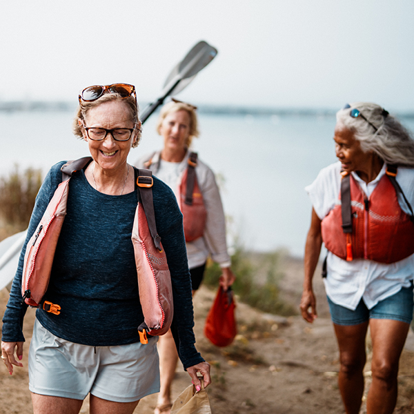 Three older women smile and laugh while carrying kayak paddles and gear along a sandy path near the water