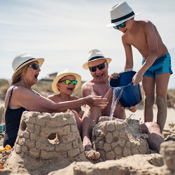 Grandparents and two grandchildren laugh together while building a sandcastle on the beach, with one child pouring water from a bucket