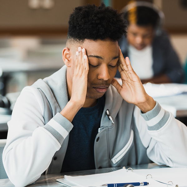 Young male sitting in a classroom with hands on temples, illustrating stress, concentration difficulties, or ringing in the ears associated with tinnitus.