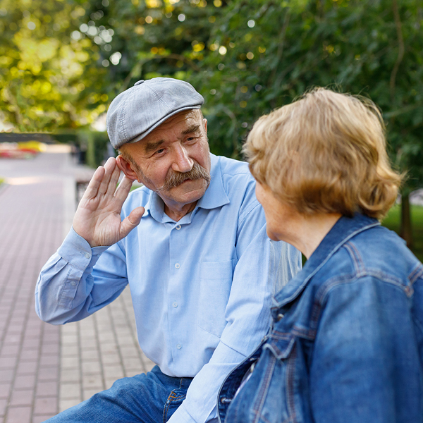 Older man cupping his hand to his ear while talking with a woman outdoors, a common sign of hearing difficulty