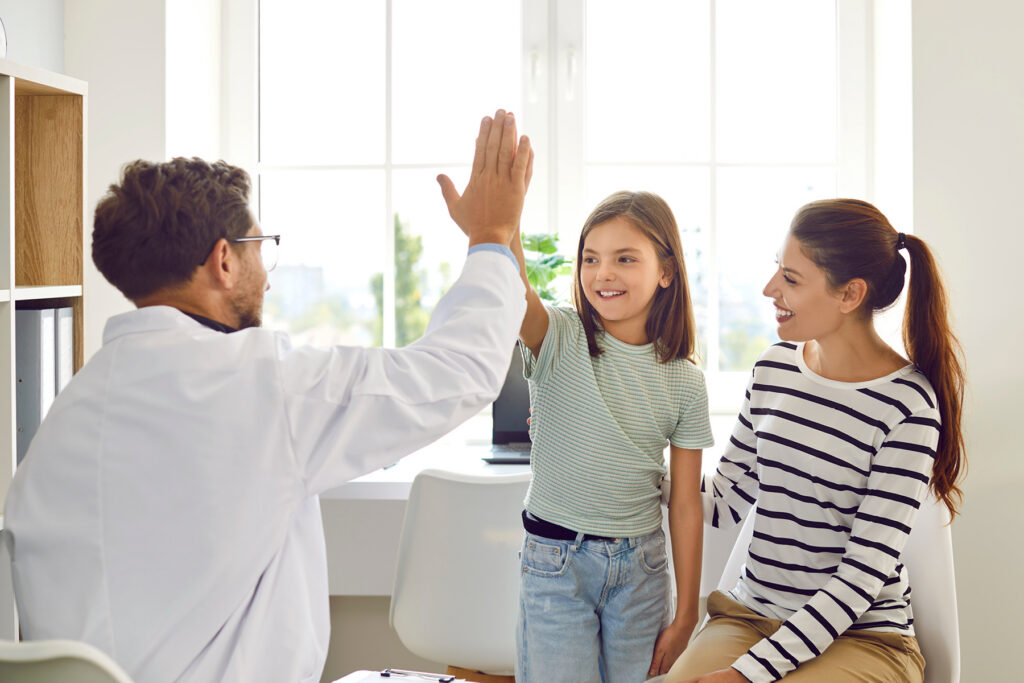 Image 3 (Hero): A smiling girl gives a high five to a healthcare provider while her mother looks on happily during a clinic visit.