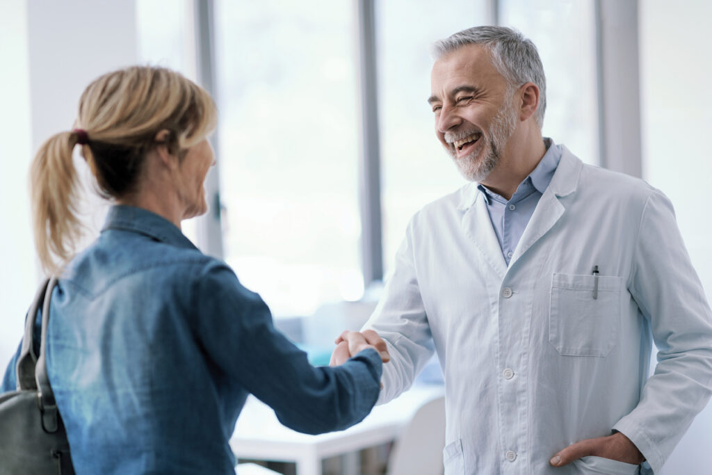 A smiling male hearing care provider in a white lab coat shakes hands warmly with a female patient in a bright clinical setting.