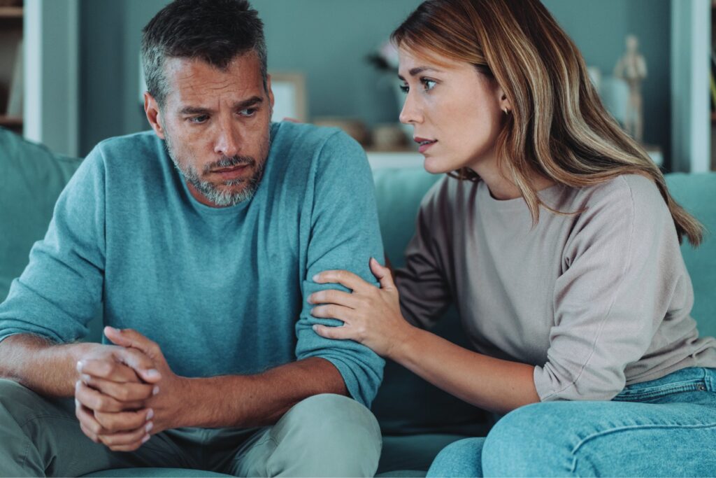 A woman speaks earnestly to a man seated beside her on a couch, her hand on his arm as he looks down with a tense expression, representing the strain that untreated hearing loss can place on close relationships