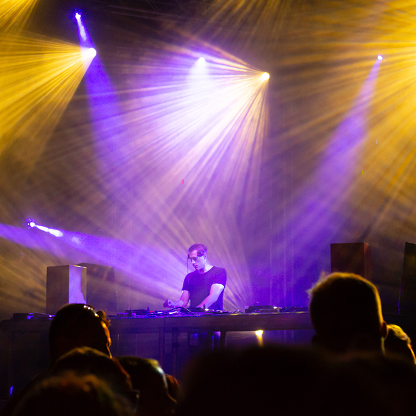 DJ performing on stage at an EDM festival with dramatic yellow and purple stage lighting beams overhead and a crowd silhouetted in the foreground