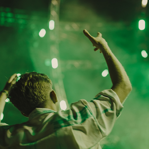 Concertgoer with arms raised at an outdoor EDM festival surrounded by green stage lighting and fog
