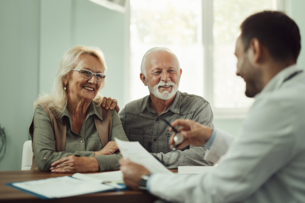 An older adult couple smiles while meeting with a healthcare provider in a clinical setting, reviewing documents together during a consultation.