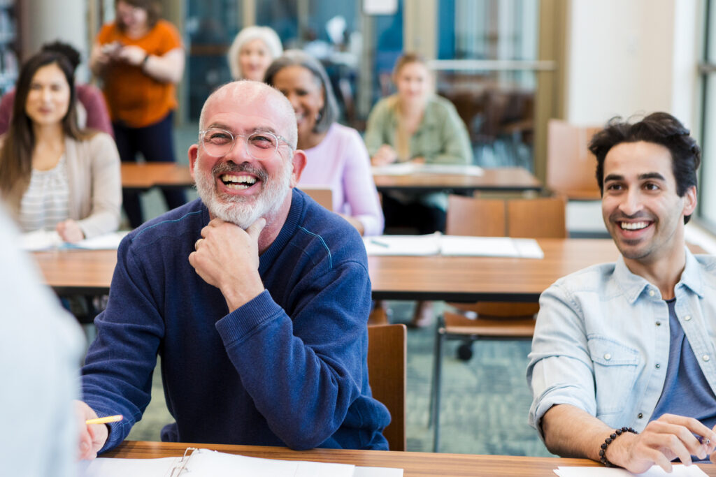 Smiling older man with glasses and a beard laughing during a group discussion in a classroom setting