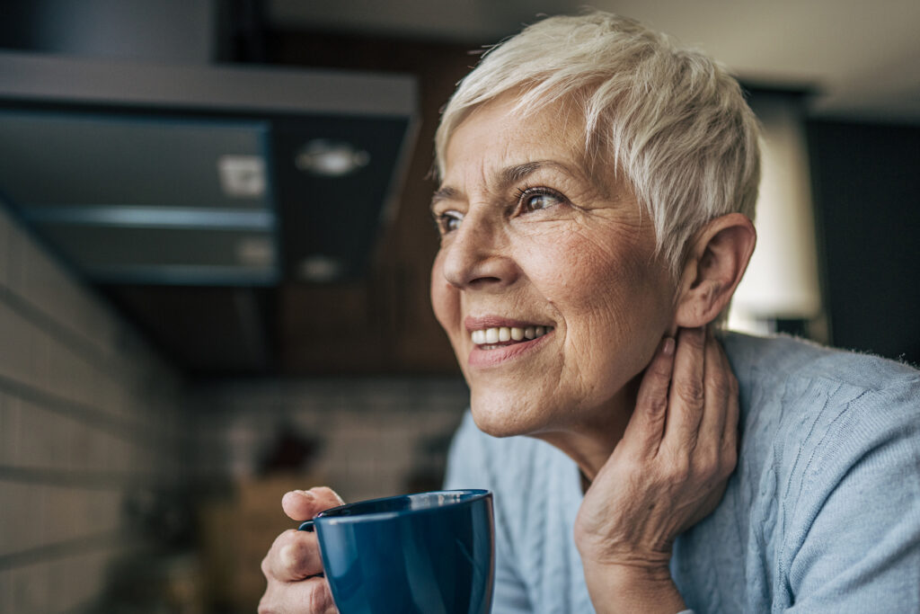 Smiling older woman with short white hair holding a blue coffee mug and looking out a window in her kitchen