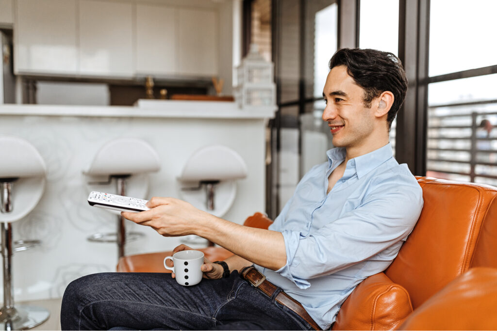 Uploaded image alt text Smiling man wearing a hearing aid relaxes in an orange leather chair with a cup of coffee while using a TV remote at home.