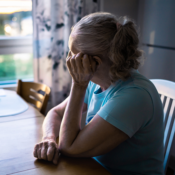 An older woman sits alone at a kitchen table, resting her head in her hand and looking out a window.