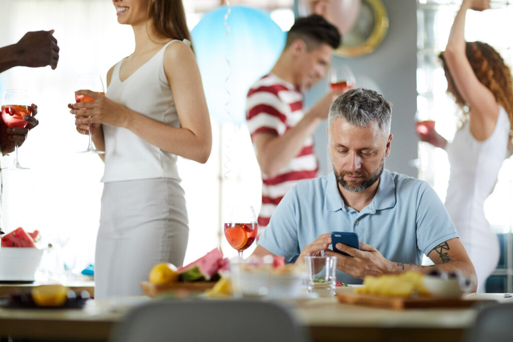 A middle-aged man with gray hair sits alone at a table looking at his phone while friends around him chat, laugh, and dance at a party.