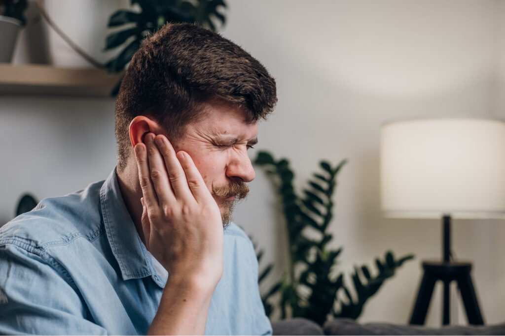 alt text for this Man at home wincing and pressing his hand against his ear, showing discomfort from sudden hearing loss or a blocked, muffled feeling in one ear.