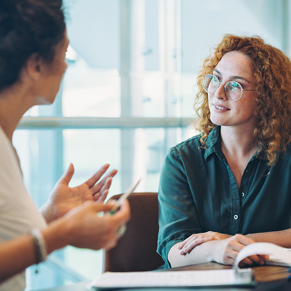 A woman with curly red hair and glasses listens attentively as another woman speaks and gestures during a conversation at a table.
