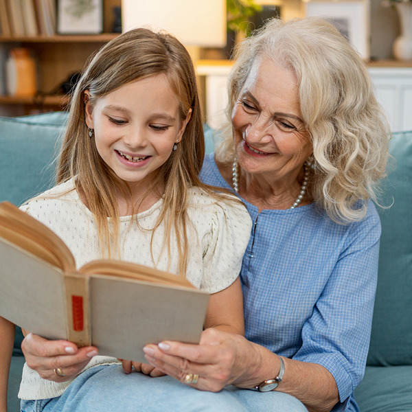 Smiling older woman with curly white hair reading a book with her young granddaughter on a living room couch.