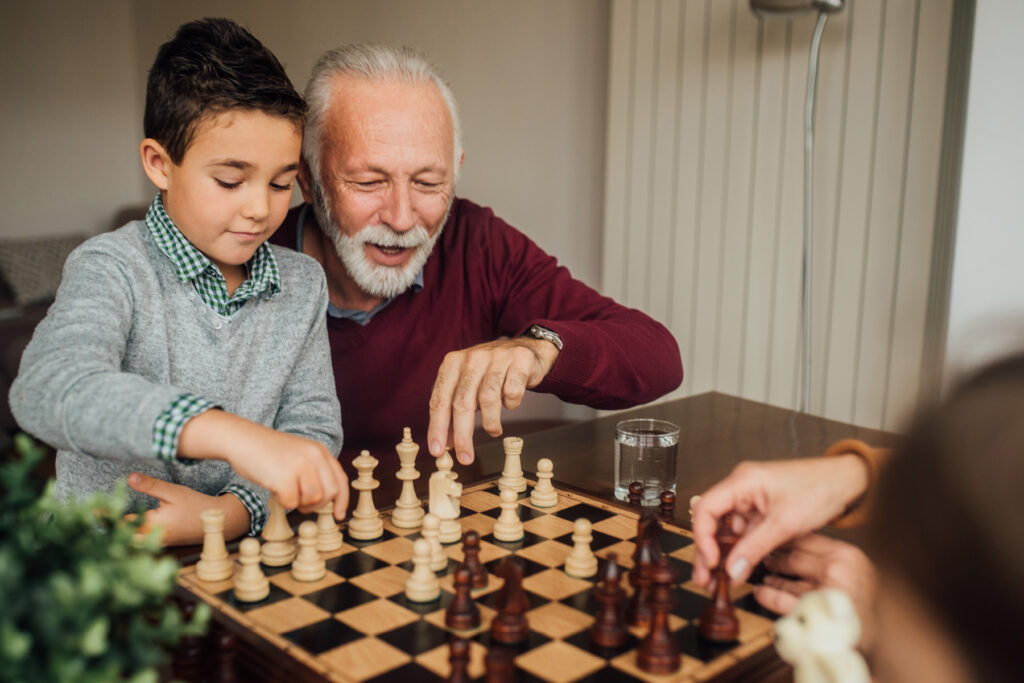 Smiling older man with a white beard playing chess at a table with his grandson.