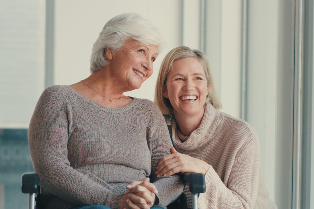 A smiling older woman sits in a wheelchair beside a laughing adult daughter who rests a hand on her arm, showing a supportive, connected relationship with a loved one who has hearing loss.