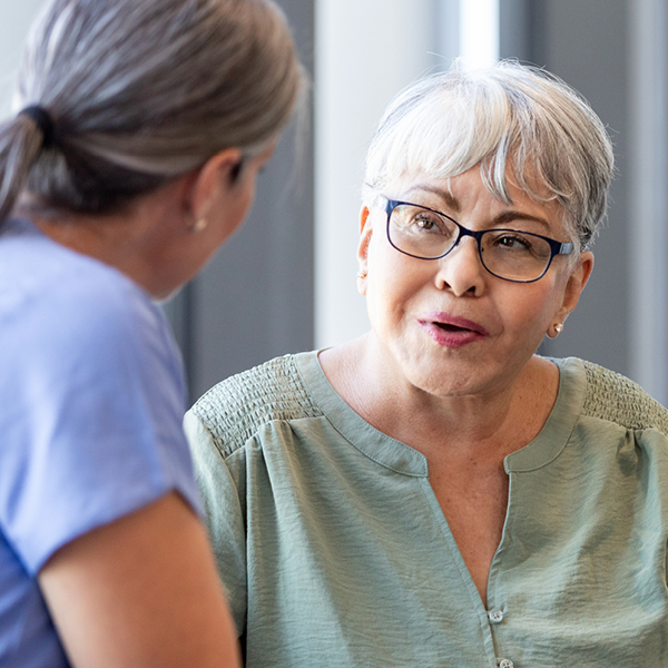 An older woman with short gray hair and glasses speaks with a woman in a blue top during a face-to-face conversation.
