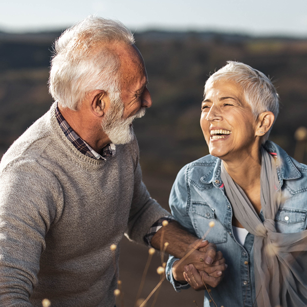 A smiling older man with a white beard walks arm in arm with a laughing older woman in a denim jacket outdoors at sunset.