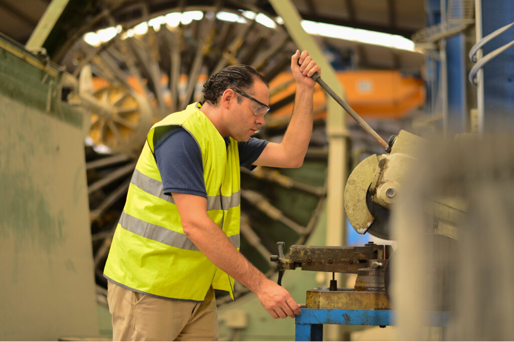 A factory worker in a high-visibility yellow vest and safety glasses pulls down the handle of a metal cutoff saw, surrounded by large industrial machinery, without wearing hearing protection.